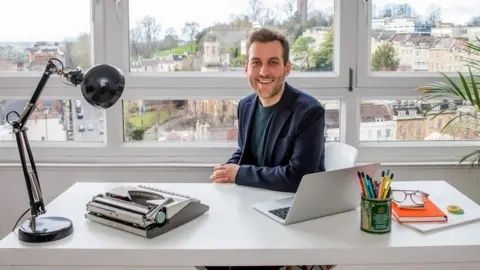 Leende man arbetar vid laptop på skrivbord med manus och pennor